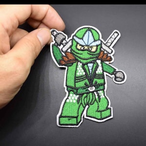 Könnte beinhalten: Gestickter Aufnäher mit einem grünen Ninja-Charakter mit weißer Umrandung. Der Ninja trägt ein grünes Outfit, eine Maske und zwei Schwerter. Der Aufnäher befindet sich auf einem schwarzen Hintergrund.