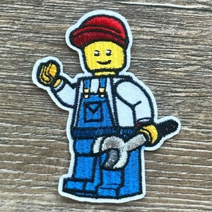 Könnte beinhalten: Gestickter Aufnäher mit einer gelben Lego-Figur, die eine rote Mütze, ein weißes Hemd und blaue Latzhosen trägt. Die Figur hält einen Schraubenschlüssel. Der Aufnäher hat einen weißen Rand.