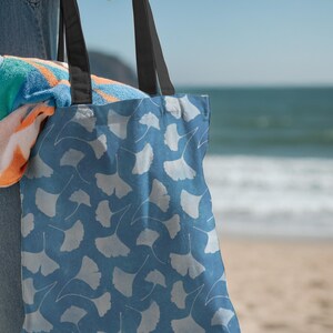 May include: A blue tote bag with a white ginkgo leaf pattern is held by a person. The bag has black straps and is filled with a colorful beach towel. The background shows the ocean and a sandy beach.