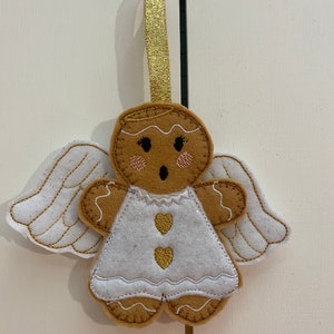May include: A gingerbread angel ornament with white wings, a white dress with gold hearts, and a gold halo. The ornament is hanging from a gold ribbon.