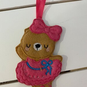 May include: A felt bear ornament in a ballerina outfit. The bear is tan with a pink bow, a pink tutu, and ballet slippers. The tutu has a blue bow and the ornament has a pink ribbon for hanging.