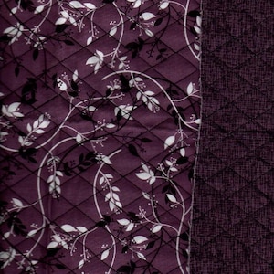 May include: Purple fabric with a white and black floral pattern. The fabric is quilted with a diamond pattern. The right side of the fabric is a solid purple with a textured pattern.