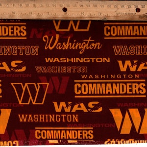 May include: Burgundy fabric printed with the Washington Commanders logo and wordmark in gold. The design includes the team name, the word "Washington", and the "W" logo. A wooden ruler, marked in centimetres, is at the top.