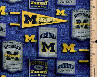 May include: Blue fabric with a repeating pattern of Michigan Wolverines logos and text. The design includes pennants, rectangular and square shapes with the letter "M" and the words "Michigan" and "Wolverines".