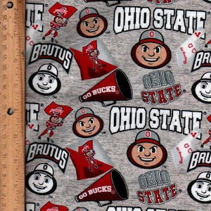 May include: Gray fabric featuring repeated patterns of Ohio State University logos, including the mascot Brutus Buckeye, the words "OHIO STATE", and red megaphones with "GO BUCKS" text. The design includes a ruler along the left edge.