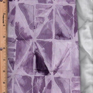 May include: Close-up of a quilted fabric with a watercolor-like design in shades of purple. The pattern features geometric shapes and is stitched with white thread in a diamond pattern. A wooden ruler is on the left side.