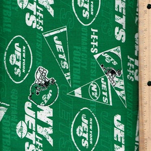May include: Green fabric with a repeating pattern of white New York Jets logos, helmets, and text. The text includes "JETS", "NEW YORK", and "J-E-T-S". A wooden ruler is on the right side of the fabric.