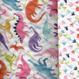 May include: White fabric with a repeating pattern of colorful dinosaurs with their names printed below. The fabric is quilted and has a white background. The dinosaurs are pink, orange, green, blue, purple, and yellow. The fabric is bordered on the right side with a white fabric with a repeating pattern of colorful dinosaur footprints.
