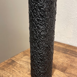 May include: A tall, cylindrical black vase with a textured, bark-like surface. The vase is made of a dark material and stands on a wooden surface. The vase has a simple, modern design.