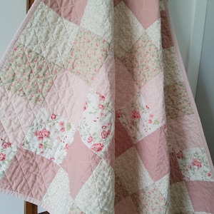 May include: A quilted blanket featuring a diamond-patterned patchwork design. The quilt consists of alternating squares in solid dusty pink and white fabric printed with small floral patterns in shades of pink, green, and beige, finished with a pink border.