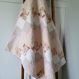 May include: A patchwork quilt featuring a diamond pattern of alternating squares. The fabric squares include solid light pink, white, beige gingham, and floral prints in shades of pink, mustard yellow, and brown. The quilt has a light pink border.