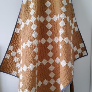 May include: A quilted blanket featuring a repeating geometric pattern of mustard yellow and white squares arranged in a diamond grid. The textile has a textured, stitched surface and is finished with a solid grey fabric binding along the edges.