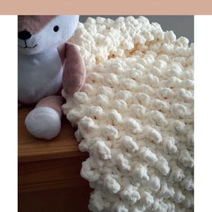Puede incluir: Una manta de bebé de crochet blanco con un diseño texturizado y abultado. La manta está drapeada sobre una superficie de madera y un juguete de peluche está sentado sobre la superficie.
