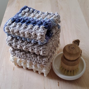 May include: A stack of four crocheted dishcloths in shades of blue and beige. The dishcloths are made with a textured stitch and have a scalloped edge. A wooden brush with a white ceramic base is in the foreground.