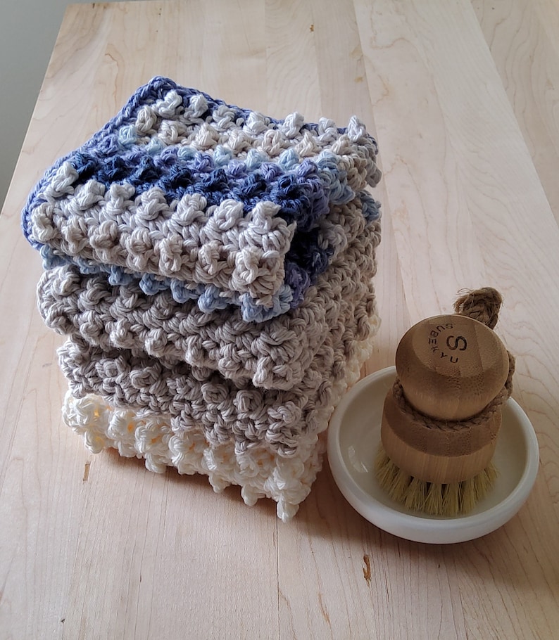 May include: A stack of four crocheted dishcloths in shades of blue and beige. The dishcloths are made with a textured stitch and have a scalloped edge. A wooden brush with a white ceramic base is in the foreground.