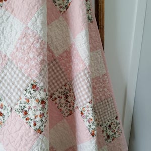 May include: A patchwork quilt featuring a repeating diamond pattern of solid light pink fabric, white fabric with a small floral print, and a beige and white gingham check fabric. The quilt has visible machine quilting stitches throughout.