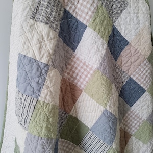 May include: A quilted blanket featuring a patchwork design of diamond-shaped fabric squares in muted tones of sage green, dusty blue, beige, and white. The fabric squares include solid colors, gingham checks, and thin stripes, all joined by stitching.
