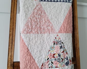 May include: A quilted blanket with a patchwork design, draped over a wooden ladder. The quilt features white, pink, and navy blue gingham fabric. Some sections have floral and rabbit patterns. The quilt is made of soft, quilted material.