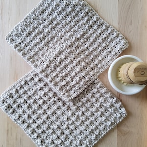 May include: Two beige crocheted dishcloths, made with a textured stitch, are stacked on a wooden surface. A white ceramic bowl with a wooden brush is in the background.