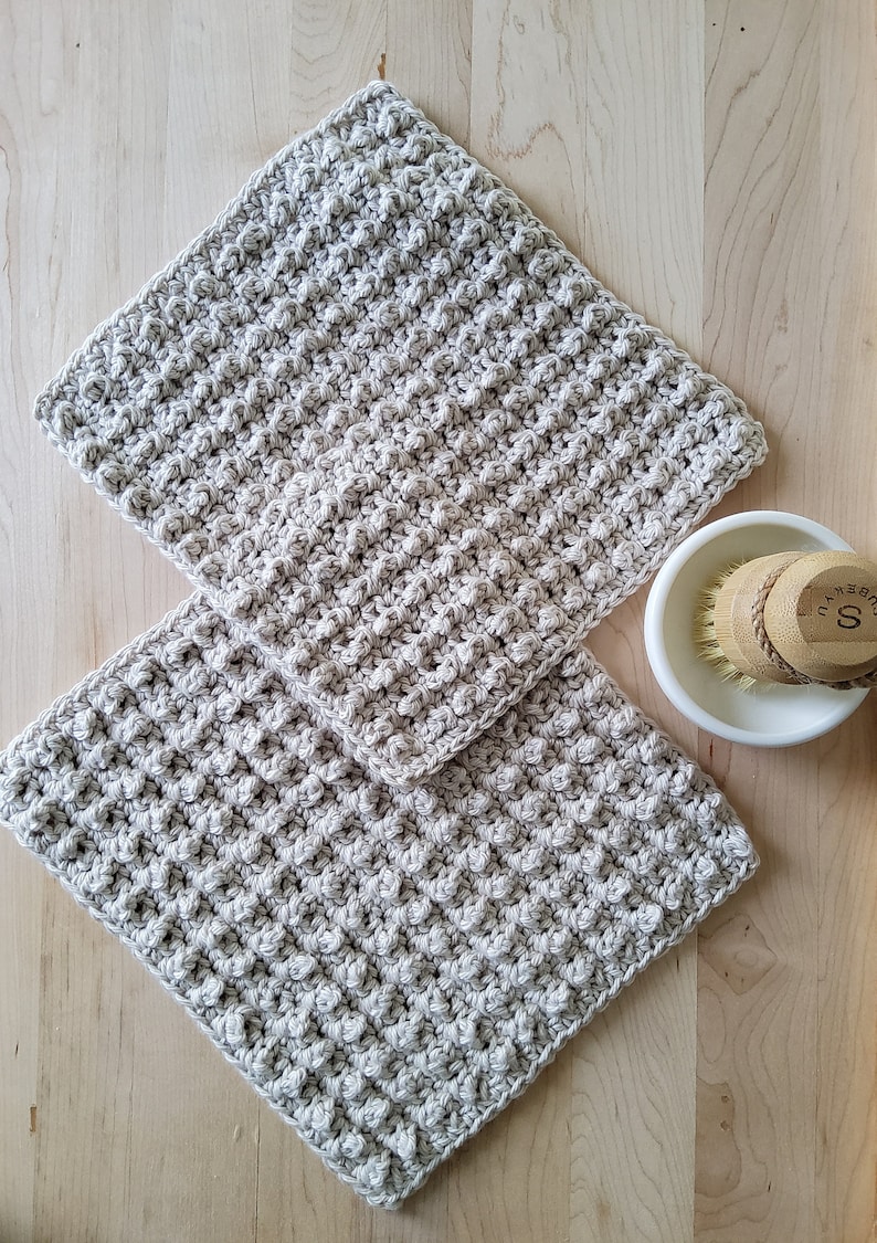 May include: Two beige crocheted dishcloths, made with a textured stitch, are stacked on a wooden surface. A white ceramic bowl with a wooden brush is in the background.