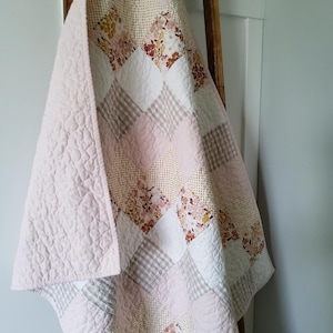 Peut inclure: Une courtepointe en patchwork présentant un motif en losange composé de carrés alternés rose pâle, blancs et beiges. Les carrés de tissu incluent des unis, des imprimés floraux et des vichys, avec un piquage texturé en forme de spirales.