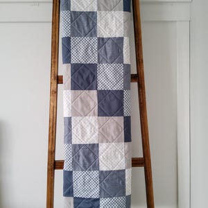 May include: A patchwork quilt with a blue, white, and gray color scheme. The quilt is hanging on a wooden ladder.