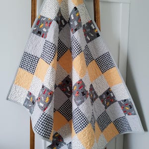 May include: A patchwork quilt with a variety of patterns, including black and white gingham, small white polka dots, and gray squares with dog illustrations. The quilt also features squares of a soft yellow color. The quilt is draped over a wooden ladder.