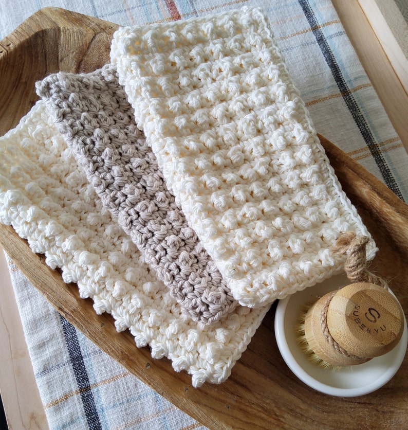 May include: Three crocheted dishcloths in white and beige, stacked on a wooden surface. The top dishcloth has a knotted handle. A wooden brush with the text "SUBEKYU" is in the background.