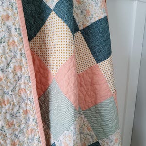 Peut inclure: Une courtepointe en patchwork composée de carrés de tissu dans des tons de bleu sarcelle, rose tendre et jaune moutarde. Le motif comprend des fleurs, des petits pois et des rayures, le tout bordé d'un biais en tissu rose uni.