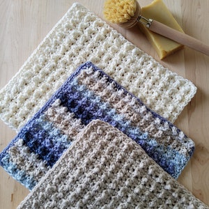 May include: Three crocheted dishcloths in different colors and patterns. The dishcloths are made with a textured stitch and are stacked on top of each other. The top dishcloth is white, the middle dishcloth is blue and beige, and the bottom dishcloth is beige.