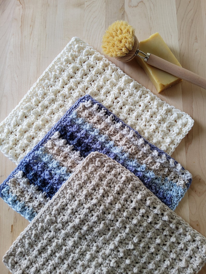 May include: Three crocheted dishcloths in different colors and patterns. The dishcloths are made with a textured stitch and are stacked on top of each other. The top dishcloth is white, the middle dishcloth is blue and beige, and the bottom dishcloth is beige.