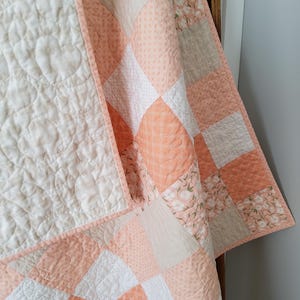 May include: A peach and white patchwork quilt with a floral pattern. The quilt is folded and draped over a wooden ladder.