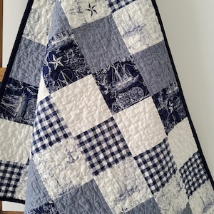 May include: A blue and white patchwork quilt with a nautical theme. The quilt features a variety of fabrics, including gingham, denim, and a white fabric with a navy blue nautical print. The quilt is quilted with a simple diamond pattern.
