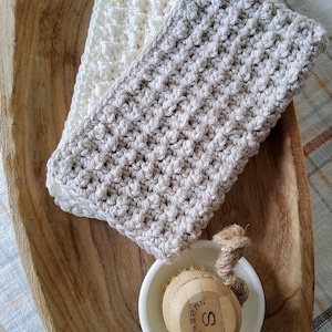 May include: Two crocheted dishcloths, one white and one beige, are displayed on a wooden tray. A small white bowl with a wooden brush is also on the tray.