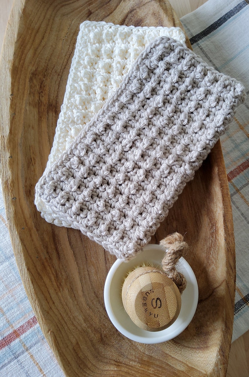 May include: Two crocheted dishcloths, one white and one beige, are displayed on a wooden tray. A small white bowl with a wooden brush is also on the tray.