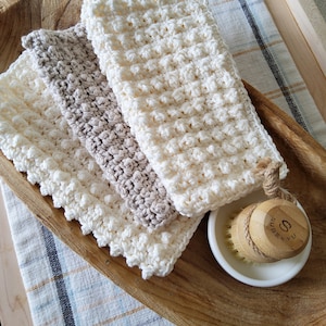 May include: Three crocheted dishcloths in white and beige, stacked on a wooden tray. A small white dish with a wooden brush is in the foreground.