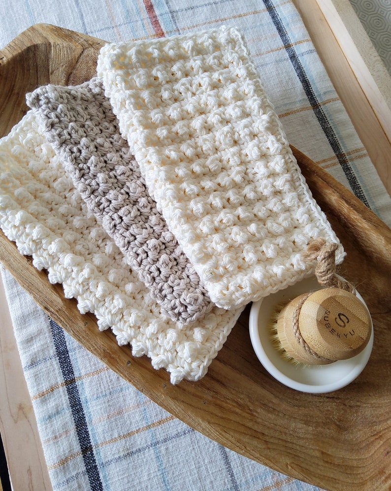 May include: Three crocheted dishcloths in white and beige, stacked on a wooden tray. A small white dish with a wooden brush is in the foreground.