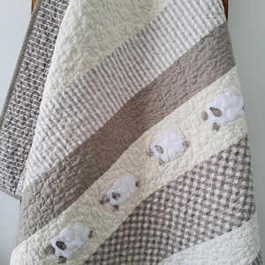 May include: A beige and white baby quilt with a diagonal pattern of gingham, stripes, and plain fabric. The quilt features embroidered white sheep appliques.