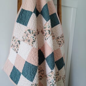 May include: A quilted blanket featuring a diamond-patterned patchwork design. The quilt consists of alternating squares in solid dusty pink, textured white, dark teal, and a floral print with green leaves and small flowers on a light background.