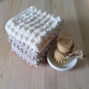 May include: Two crocheted dishcloths, one white and one beige, stacked on top of each other. A wooden brush with a white ceramic dish is next to the cloths.