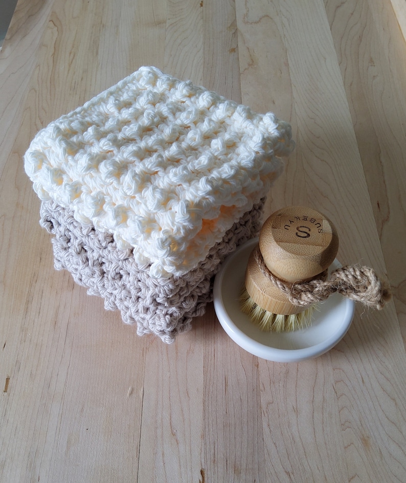 May include: Two crocheted dishcloths, one white and one beige, stacked on top of each other. A wooden brush with a white ceramic dish is next to the cloths.