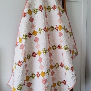 May include: A patchwork quilt featuring a repeating diamond pattern of white squares interspersed with smaller squares in shades of dusty pink, mustard yellow, olive green gingham, and a floral print in deep red, finished with a solid pink border.