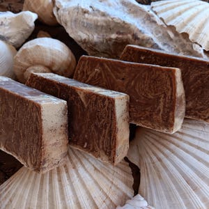 May include: Four bars of handmade soap with a brown and white swirl pattern. The soap is displayed on a bed of seashells.