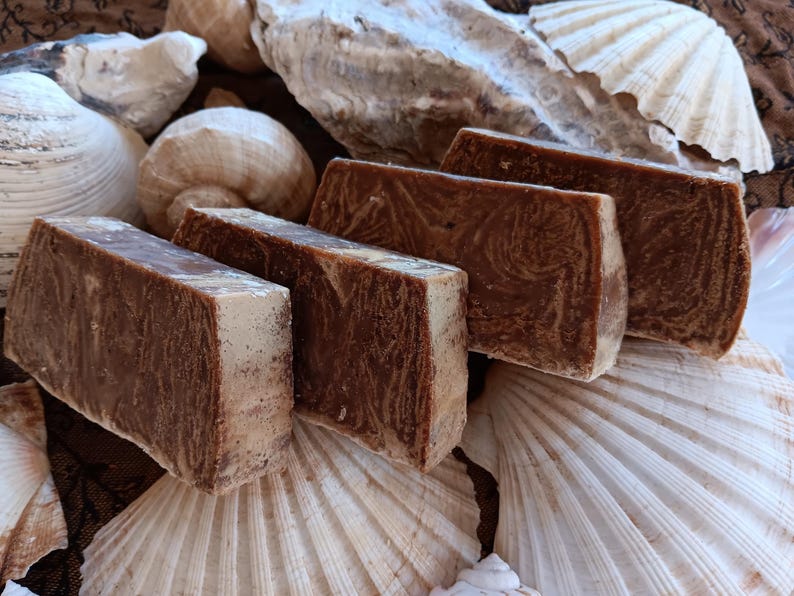 May include: Four bars of handmade soap with a brown and white swirl pattern. The soap is displayed on a bed of seashells.