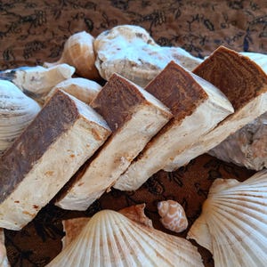 May include: Four bars of brown soap stacked on top of each other. The soap is a light brown colour with darker brown streaks. The soap is sitting on a brown fabric background with seashells.