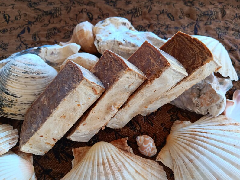 May include: Four bars of brown soap stacked on top of each other. The soap is a light brown colour with darker brown streaks. The soap is sitting on a brown fabric background with seashells.