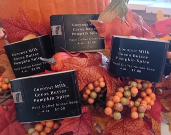 Könnte beinhalten: Vier handgefertigte Seifenstücke mit schwarzen Etiketten, mit dem Text "Coconut Milk Cocoa Butter Pumpkin Spice". Die Seifen wiegen jeweils 113 g. Die Seifen sind von Herbstlaub umgeben, mit einem Kürbis im Hintergrund.