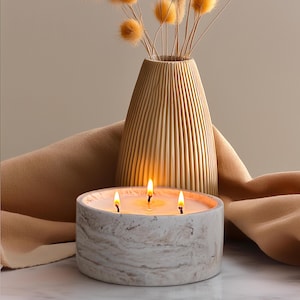 May include: A white marble candle with three wicks burning. The candle is sitting on a brown fabric surface. A vase with dried flowers is in the background.