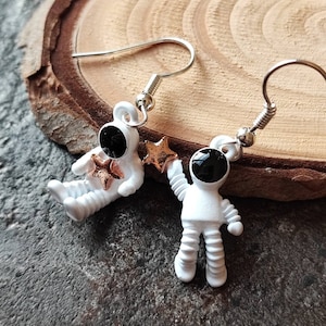 Può includere: Un paio di orecchini a forma di astronauta argentati con un astronauta bianco che tiene una stella dorata. L'astronauta ha una maschera facciale nera.