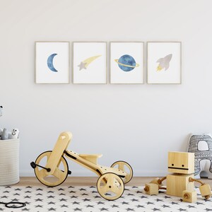 May include: A wooden tricycle with a light wood finish and black wheels. The tricycle is on a white and gray star patterned rug. There are three framed prints on the wall behind the tricycle. The prints are of a crescent moon, a shooting star, a planet with rings, and a rocket ship. All of the prints are in shades of blue and yellow.